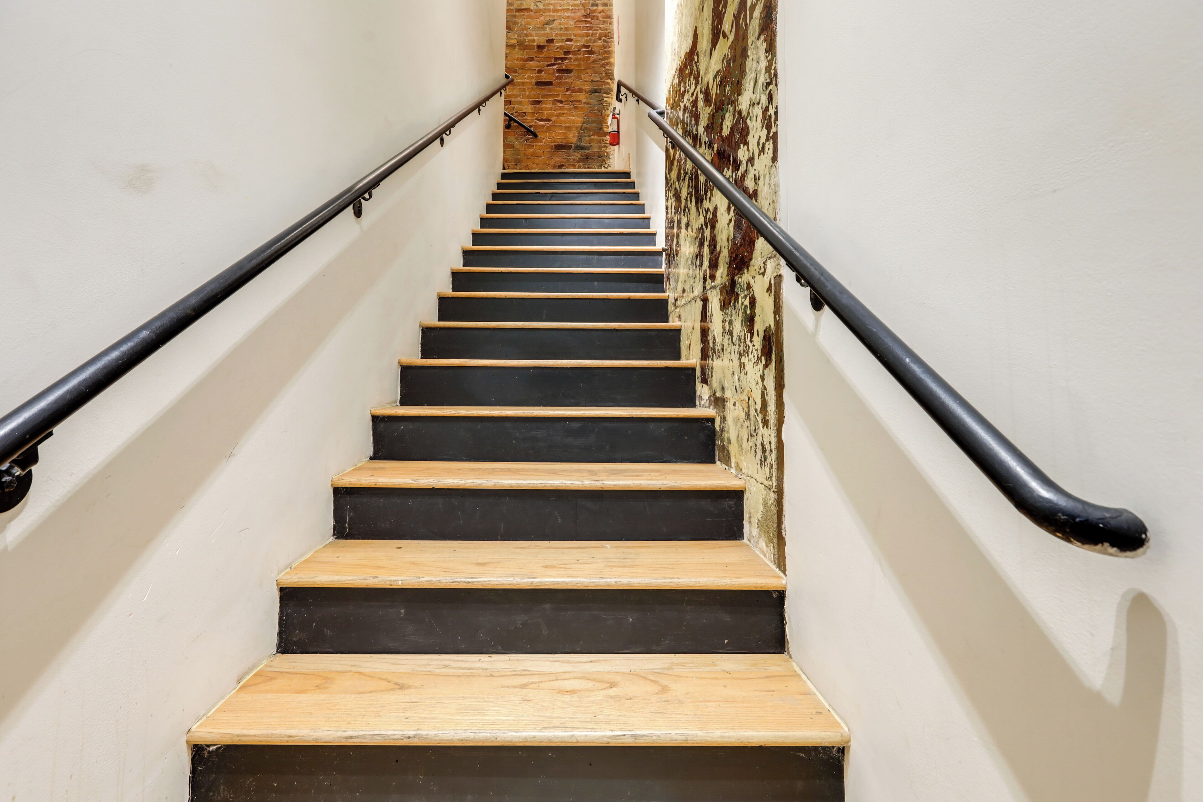A staircase with wooden steps and black risers, flanked by white walls and a brick wall at the top.