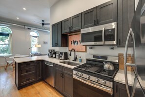 A modern kitchen with dark cabinetry, stainless steel appliances, a large island with a sink, and a view of the outdoors through a window. - Walkable & Stylish! Cincy Loft in Prime OTR Spot (Cincinnati)