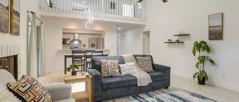 A spacious living room with a grey sofa, a coffee table, and a patterned rug. There is a balcony with a white railing and a ceiling fan.