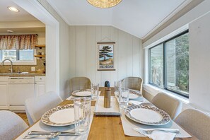Comedor con una mesa de madera dispuesta para cuatro personas, sillas de mimbre alrededor y una cocina al fondo, con vista por la ventana.