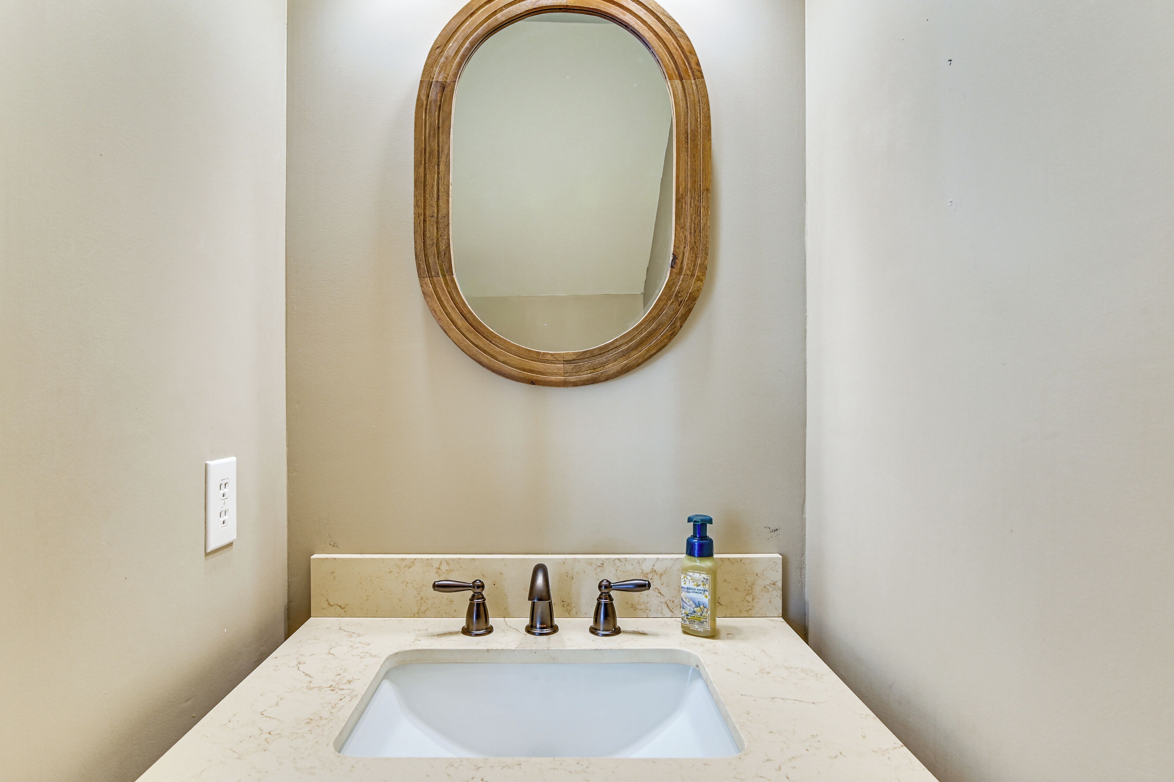 A bathroom with a white sink, two faucets, a mirror with a wooden frame, and a bottle of hand soap on the counter.