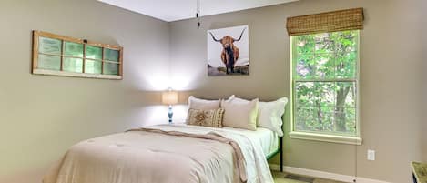 Un dormitorio con cama, ventilador de techo, ventana con vista a árboles y un cuadro de vaca colgado en la pared.