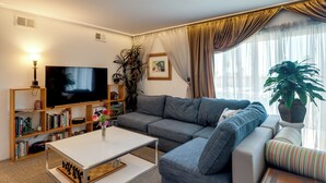 A living room with a television, sofa, coffee table, bookshelf, and a large window with curtains. - Walkable & Renovated Gem! Only 8 Mi to Dtwn Tucson (Tucson)
