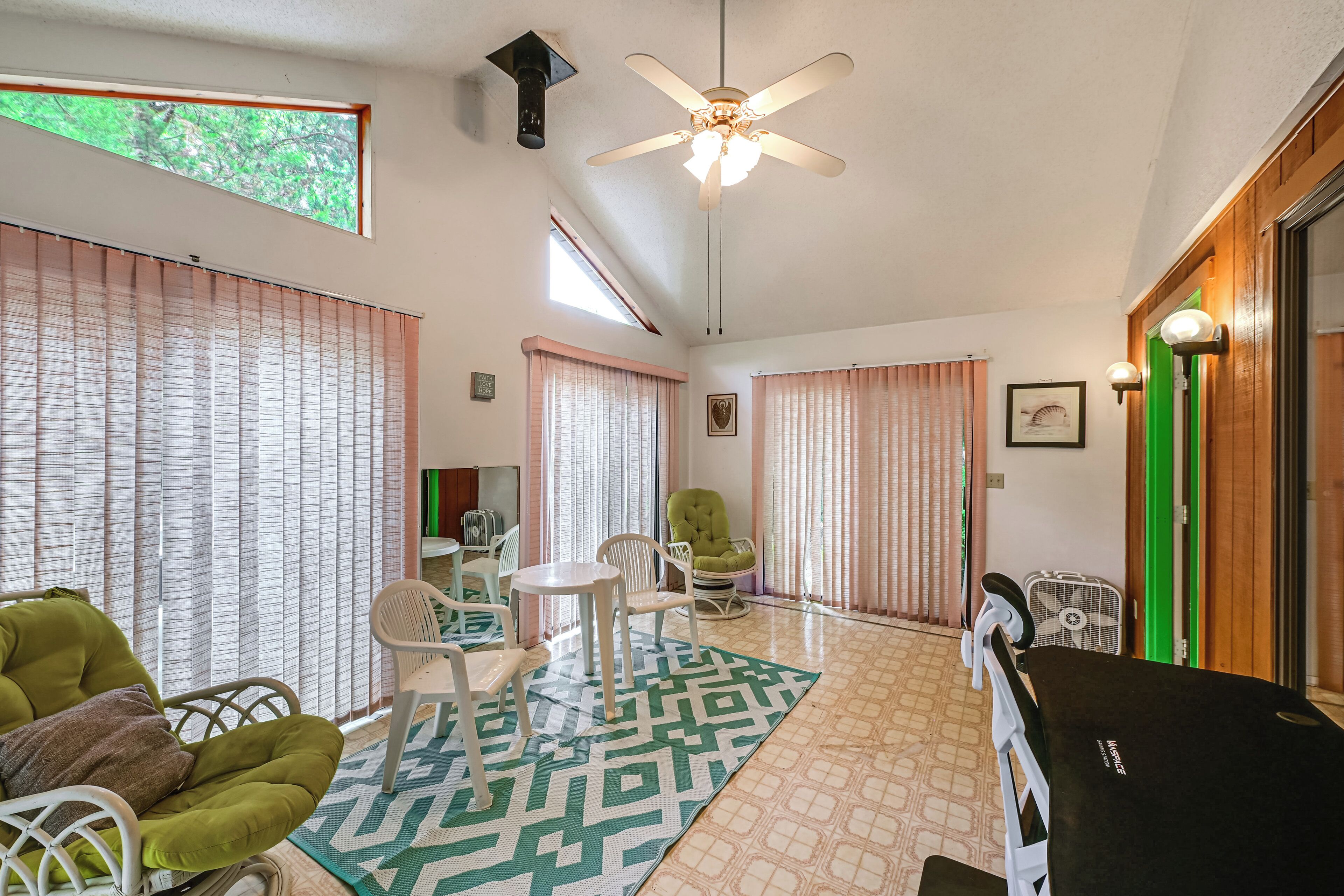 A room with a ceiling fan, a patterned rug, a green chair, a white chair, and a small table.