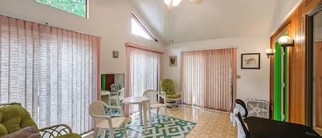 A room with a ceiling fan, a patterned rug, a green chair, a white chair, and a small table.