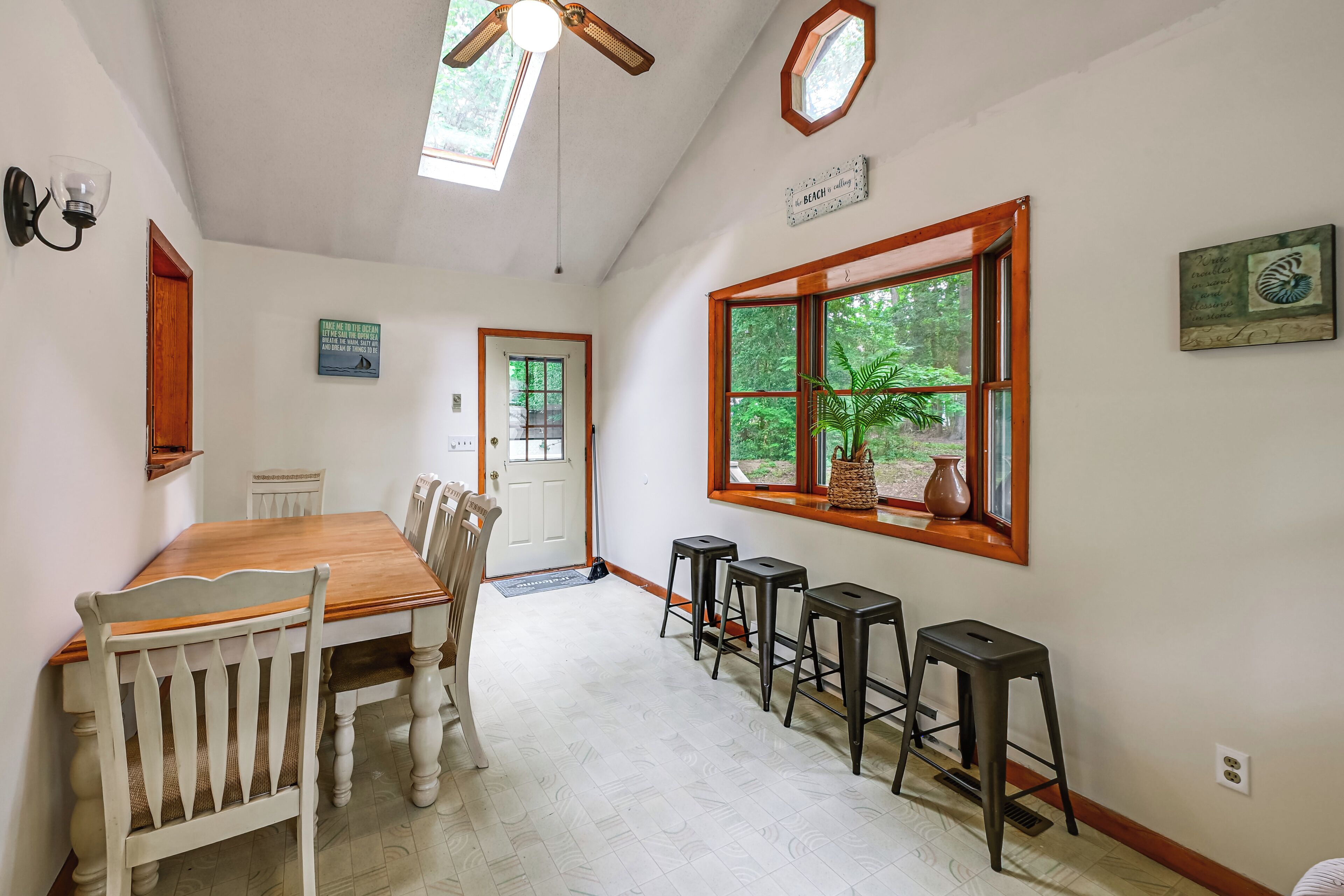 Uma sala de jantar com mesa de madeira e cadeiras brancas, ventilador de teto e janela com vista para o verde.