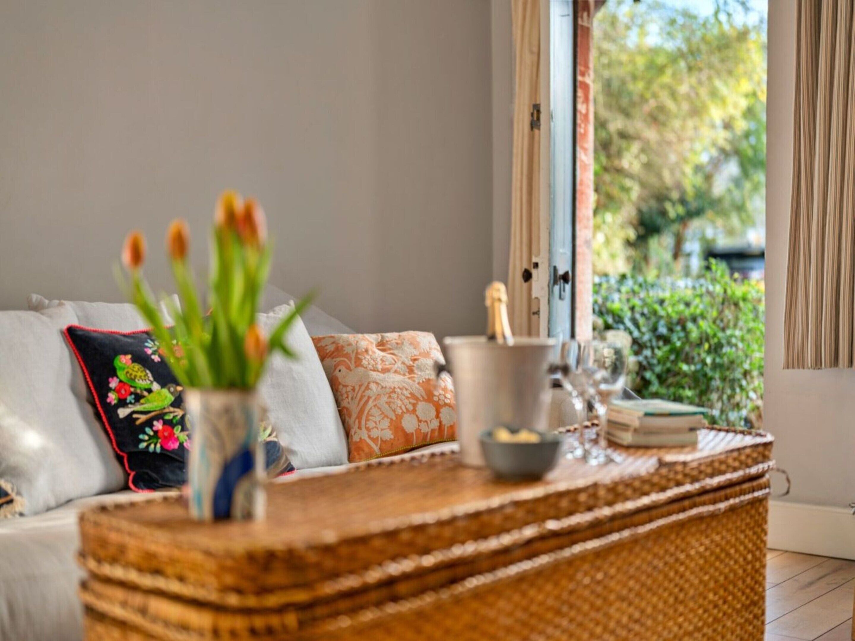 Une pièce comprenant un canapé, une table d’appoint en osier, un vase avec des fleurs et une fenêtre donnant sur un paysage verdoyant.
