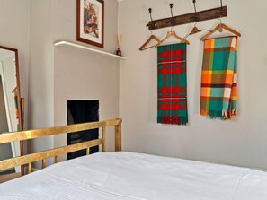 A bedroom with a wooden bed frame, a white mattress, a fireplace, and two colorful blankets hanging on a wooden rack. - Historic Wiltshire Garden Cottage (Trowbridge)