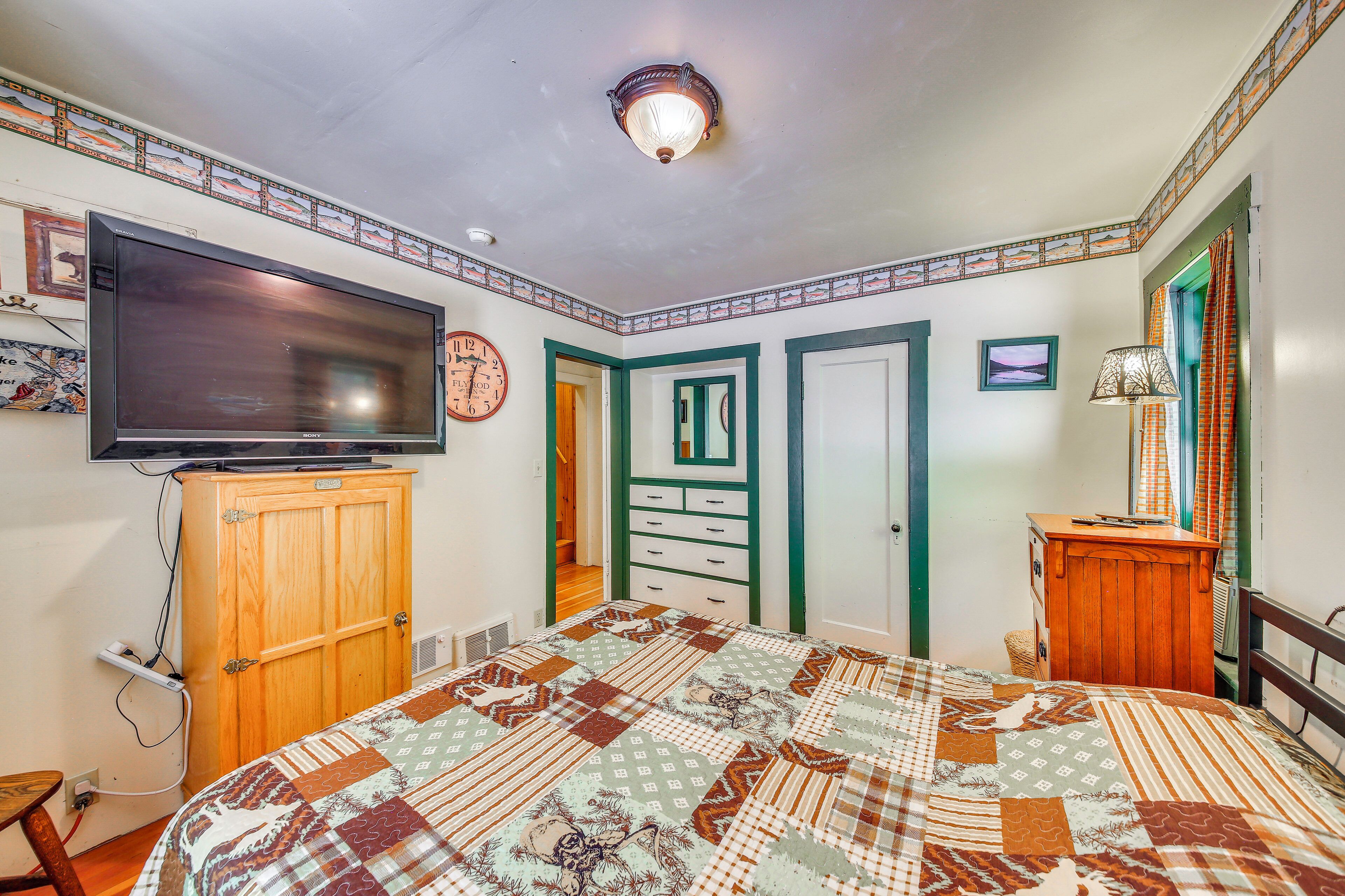 A bedroom with a large flat-screen TV, a wooden cabinet, a quilted bedspread, and a wall clock.