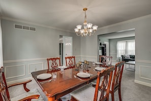 A dining room with a wooden table set for six, surrounded by chairs, a chandelier, and a view into a living area.