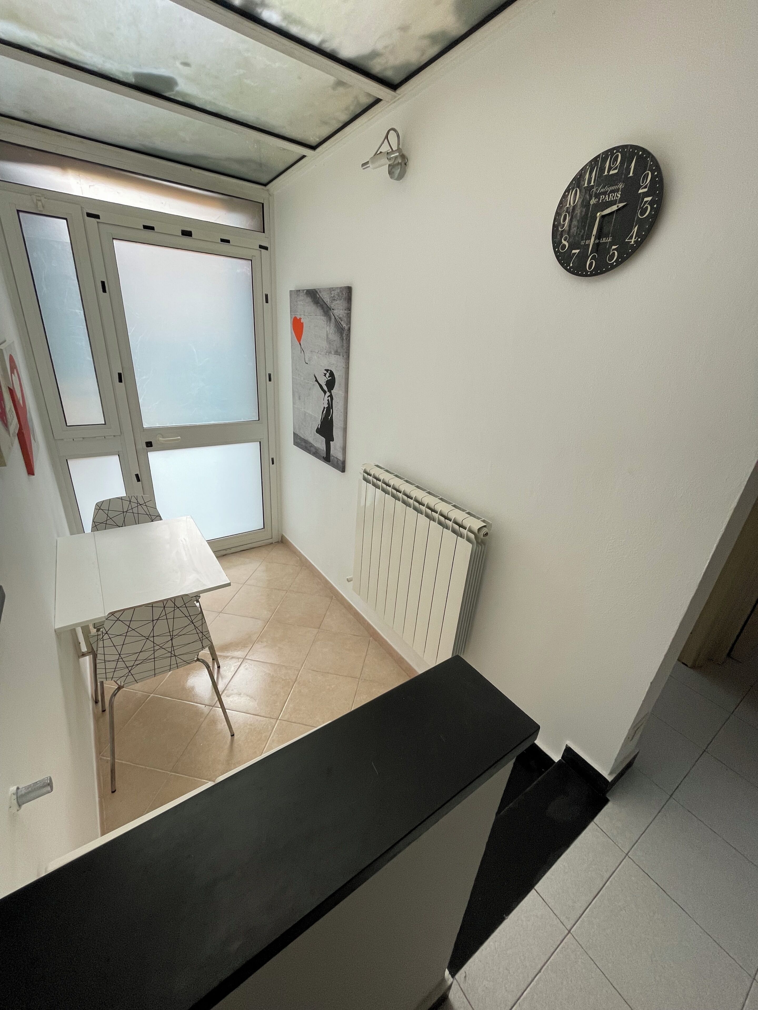 A room with a radiator, a wall clock, a table with a chair, and a window with frosted glass panels.