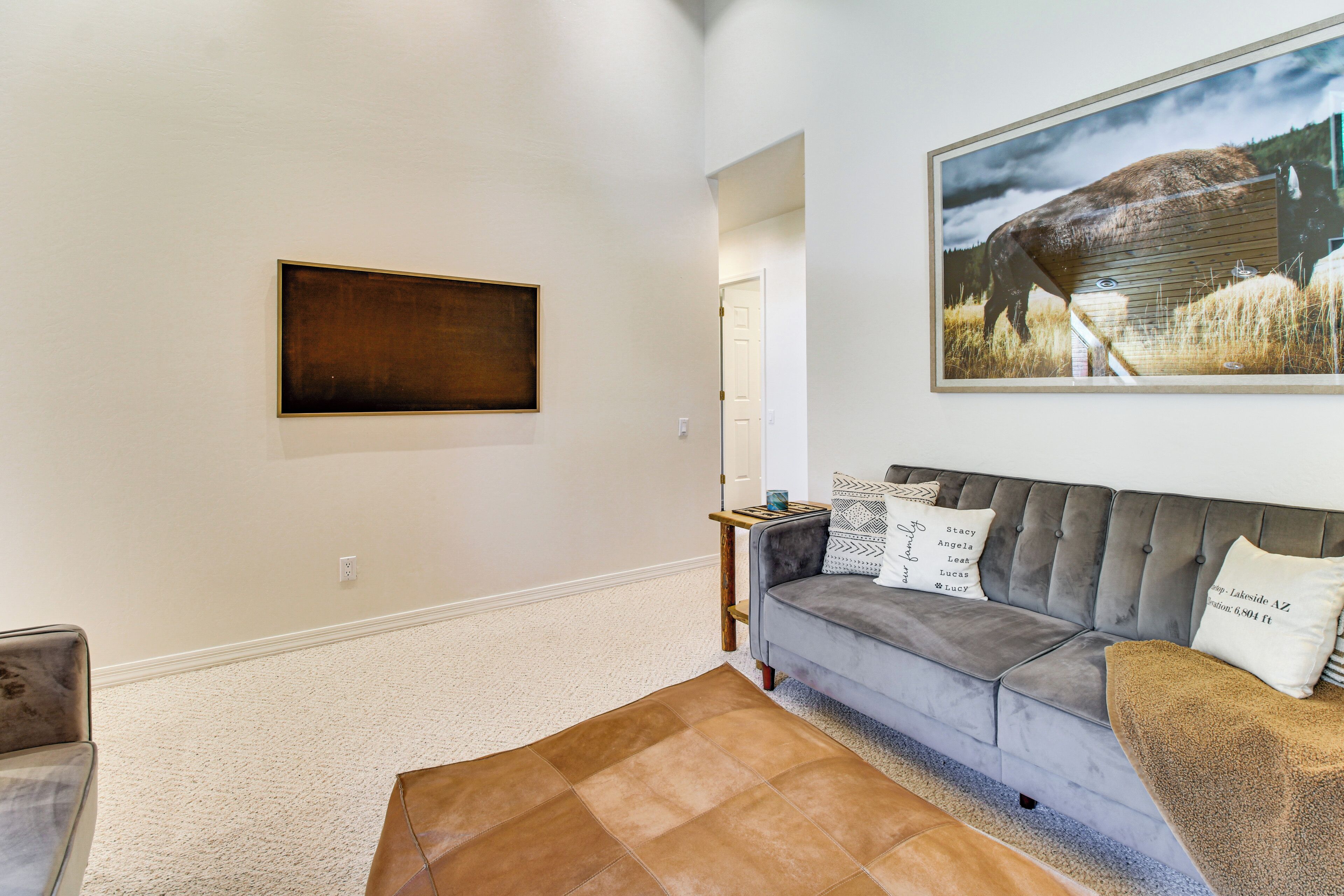 A living room with a grey sofa, a brown leather ottoman, a framed picture on the wall, and a small table with a candle.