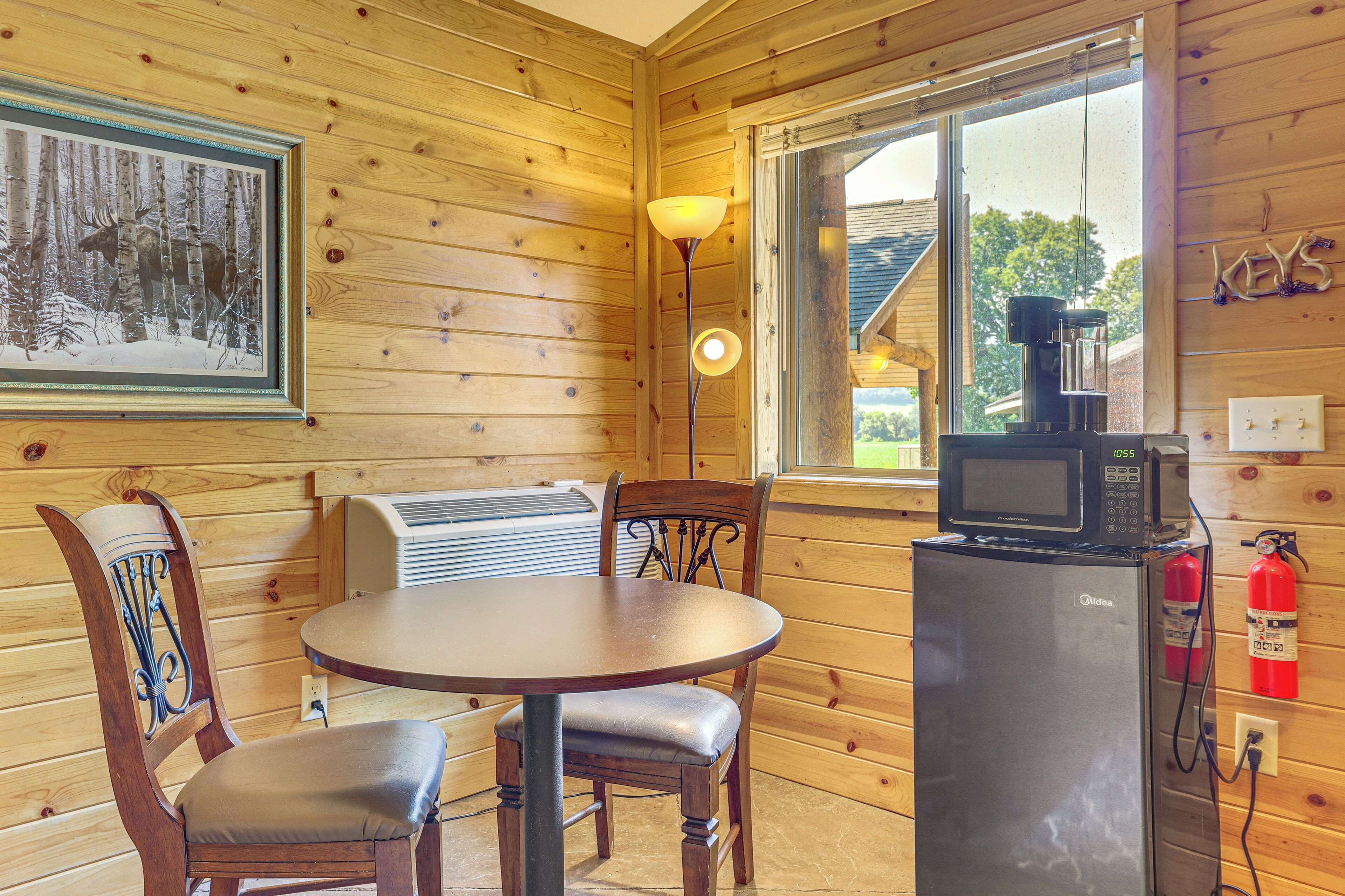A wooden cabin interior with a dining table, chairs, a ceiling fan, a mounted television, and a window with a view of a house and trees.