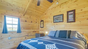 A wooden cabin room with a bed, a window with curtains, and wall-mounted pictures.