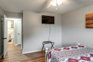 Un dormitorio con una cama, televisor montado, un cuadro y una puerta que da a un baño.