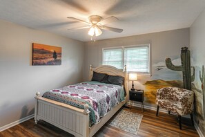 A bedroom with a bed, a ceiling fan, a window with blinds, a nightstand, a lamp, a chair, and a painting on the wall.