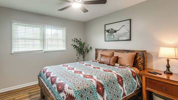A bedroom with a bed, a ceiling fan, a window with blinds, a plant, a framed picture on the wall, and a nightstand with a lamp.