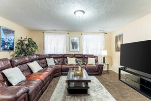 Ein Wohnzimmer mit Ecksofa, Couchtisch, Fernseher und zwei mit Vorhängen versehenen Fenstern.