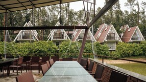 Un espacio comedor al aire libre techado, con mesas de vidrio y sillas de mimbre, con vistas a una hilera de edificios con techos triangulares.