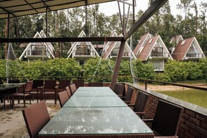 Un espacio comedor al aire libre techado, con mesas de vidrio y sillas de mimbre, con vistas a una hilera de edificios con techos triangulares.