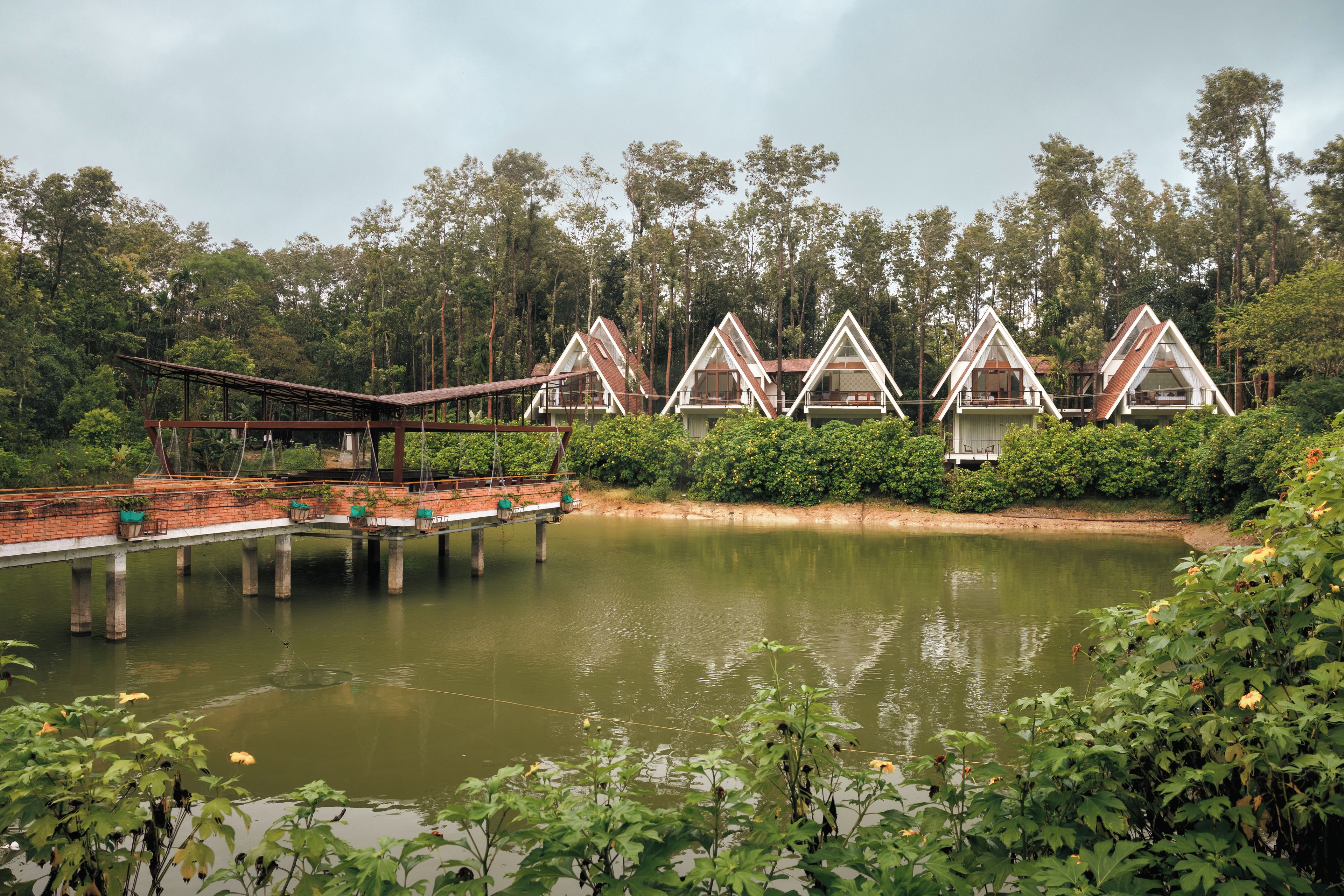 A row of triangular-roofed cabins with a bridge leading to them, set in a forested area with a body of water in the foreground.