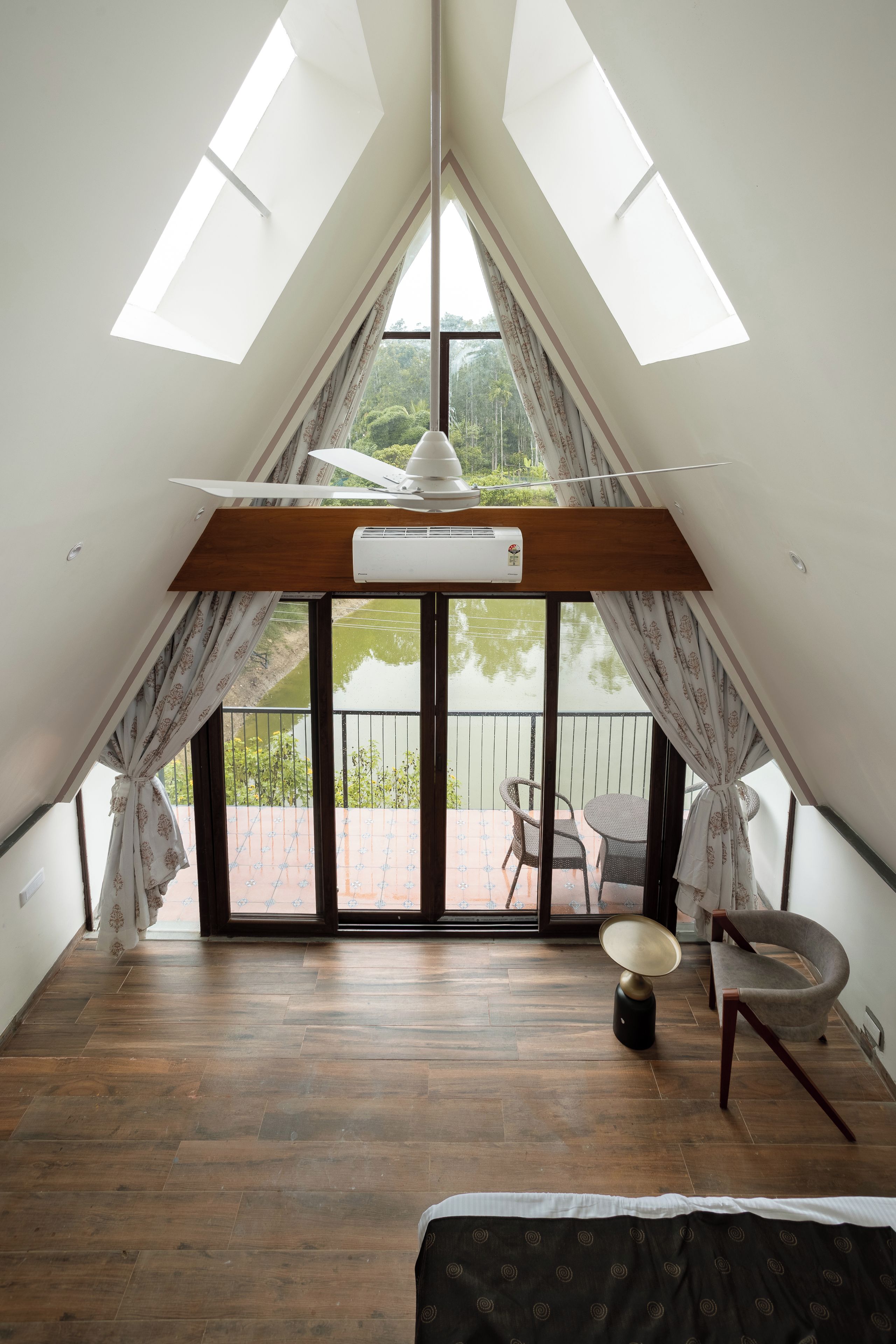 Habitació amb sostre inclinat, lluminàries al sostre, ventilador de sostre, aparell d'aire condicionat, finestra amb cortines i vista a un balcó amb mobles d'exterior, així com un edredó estampat.