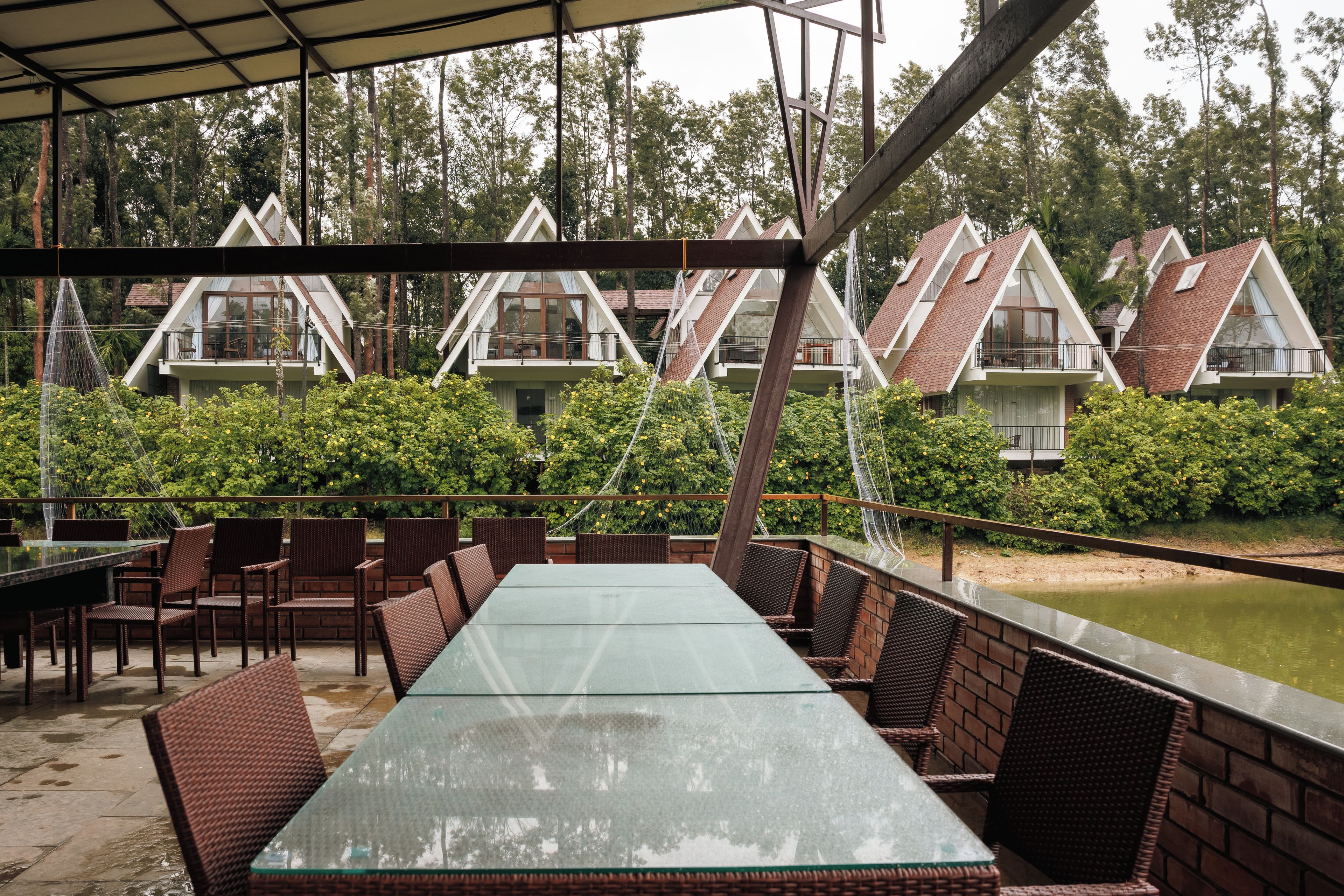 A covered outdoor dining area with wicker furniture and a glass table, surrounded by greenery and a water feature.