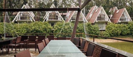 A covered outdoor dining area with wicker furniture and a glass table, surrounded by greenery and a water feature.