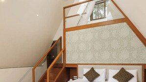 Un dormitorio con techo inclinado, cama con estructura de madera, papel tapiz estampado y vista al exterior por una ventana.