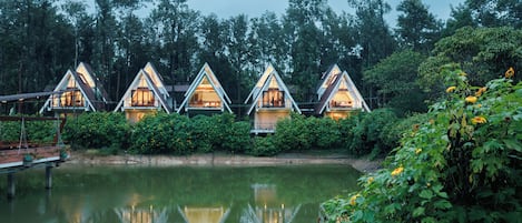 A row of triangular-roofed cabins with large windows, situated next to a calm body of water, surrounded by dense greenery.