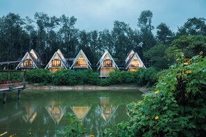 Une rangée de cabanes au toit triangulaire, dotées de grandes fenêtres, est située au bord d’une étendue d’eau calme, entourée d’une végétation luxuriante.
