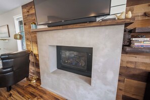 Ein modernes Wohnzimmer mit einem Flachbildfernseher über einem Kamin, einem Ledersessel und einem Bücherregal voller Bücher.