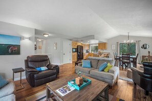 Ein geräumiges Wohnzimmer mit Ledersessel, grauem Sofa mit bunten Kissen, Couchtisch aus Holz mit einer Zeitschrift und einer Küche im Hintergrund.