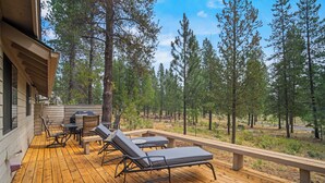 Um deck de madeira com móveis de exterior, incluindo espreguiçadeiras e uma mesa, com vista para uma área arborizada.