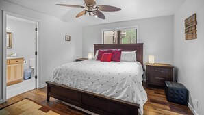 Quarto com cama, mesas de cabeceira, ventoinha no teto, janela e uma obra de arte na parede.