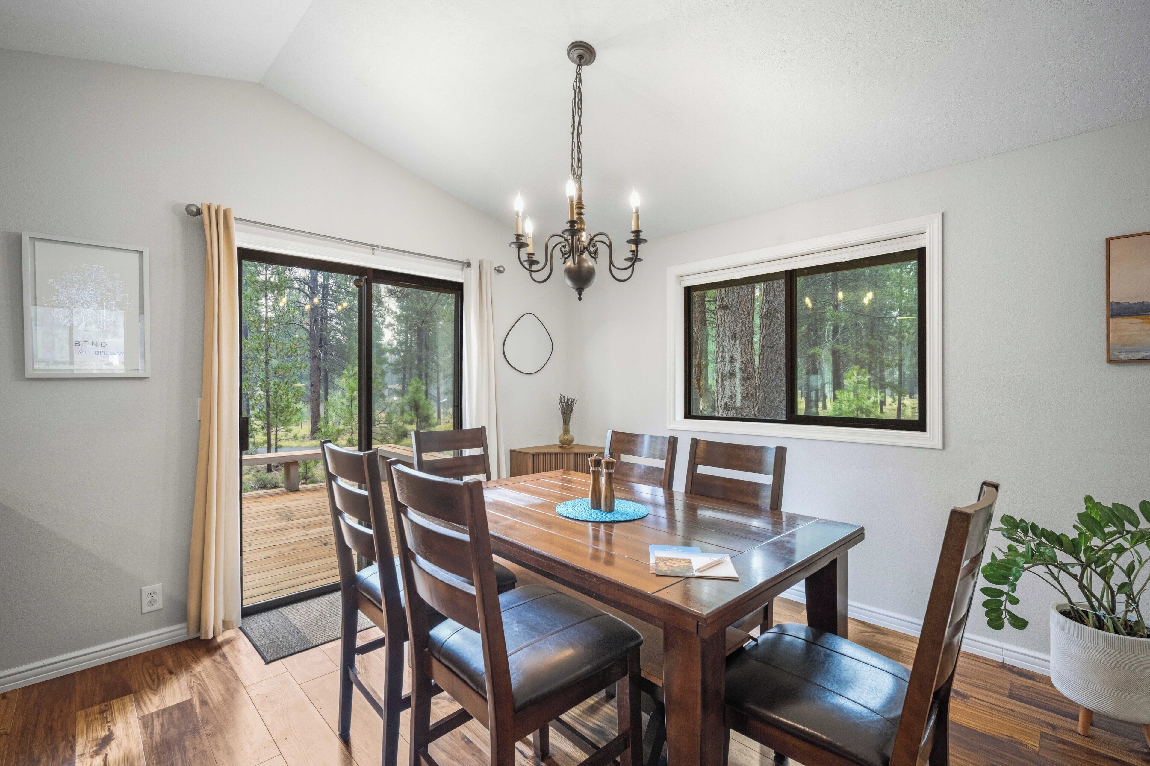 Comedor con mesa de madera, sillas, una lámpara de araña y grandes ventanales con vistas al bosque.