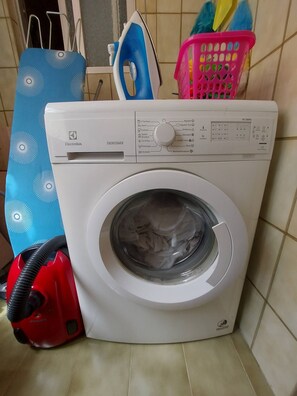 A front-loading washing machine with a control panel, a red vacuum cleaner, and a blue ironing board in a laundry area. - SPACIOUS APARTMENT 100 METERS FROM THE BEACH IN NERJA (Nerja)