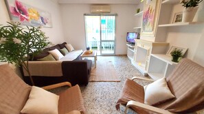 Ein Wohnzimmer mit einer Couch, Sesseln, einem Couchtisch und einem Fernseher. Eine Pflanze, ein Gemälde und ein Regal mit Dekorationen sind ebenfalls vorhanden.