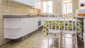 Een keuken met witte kasten, een granieten aanrecht, een eettafel met een tafelkleed met een fruitprint, en een raam met een metalen traliewerk.