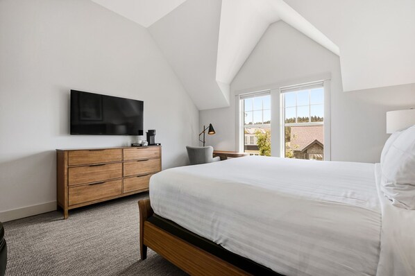 A bedroom with a large bed, a flat-screen TV mounted on the wall, a wooden dresser, a nightstand with a lamp, and a window with a view of a house.