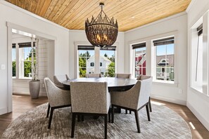A dining area with a round table, six chairs, a chandelier, and large windows with a view of buildings.