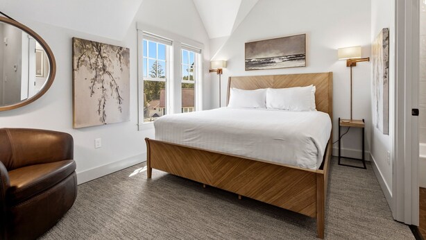 Un dormitorio con una cama de madera, un sillón de cuero marrón, un ventanal grande y obras de arte en las paredes.