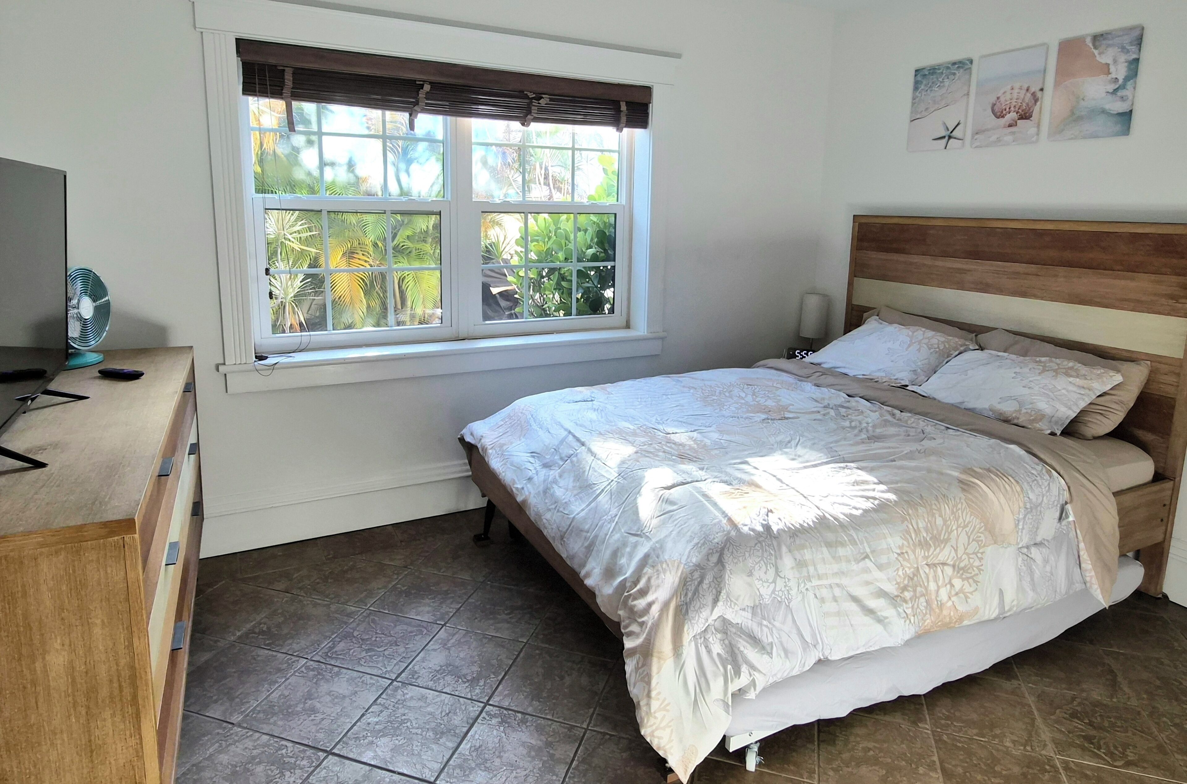 Un dormitorio con cama, ventana con persianas, lámpara de noche y ventilador sobre un tocador.