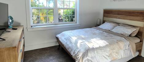A bedroom with a bed, a window with blinds, a bedside lamp, and a fan on a dresser.