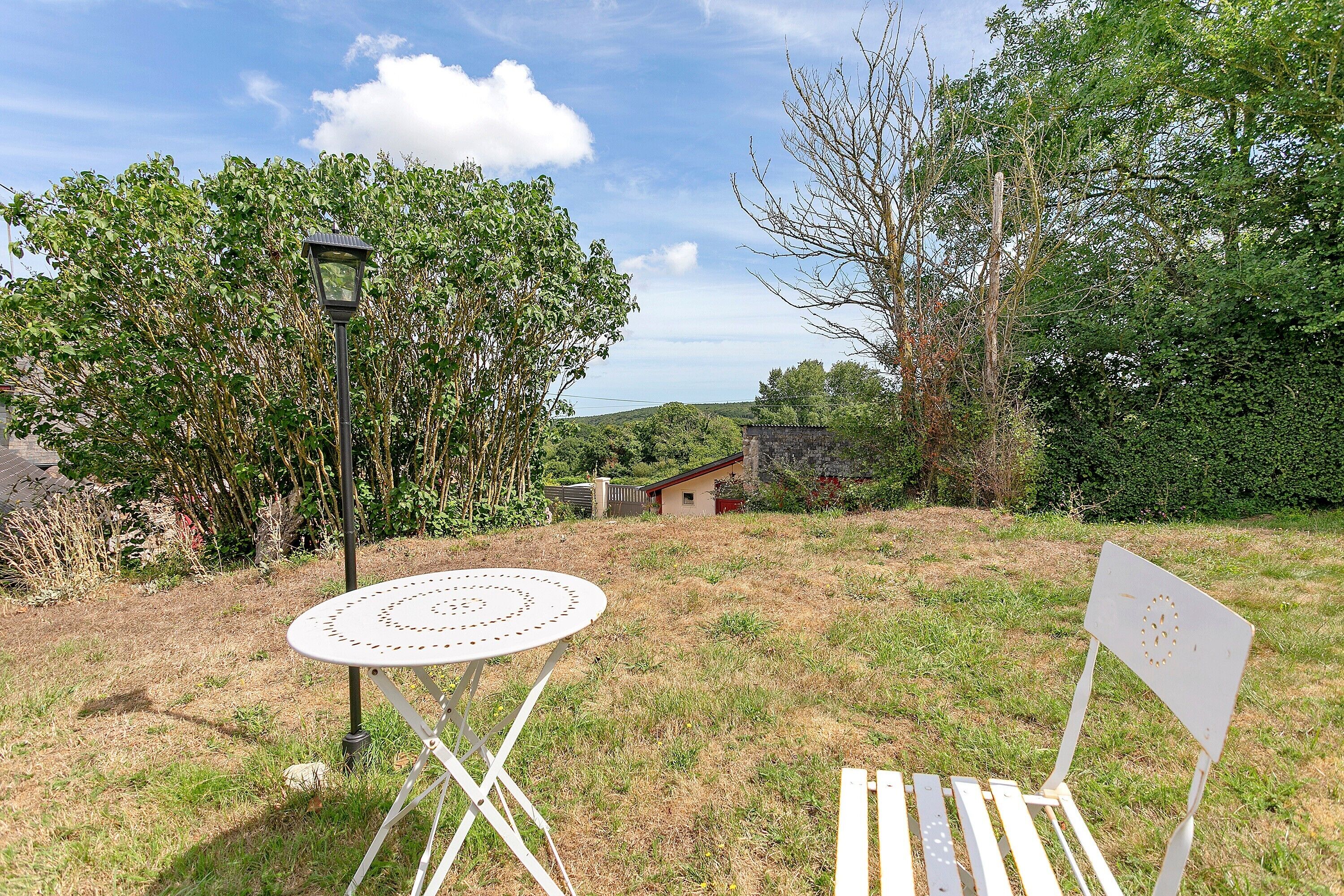 An outdoor seating area with a table and chairs, a lamp post, and a view of a rural landscape with trees and a stone building.