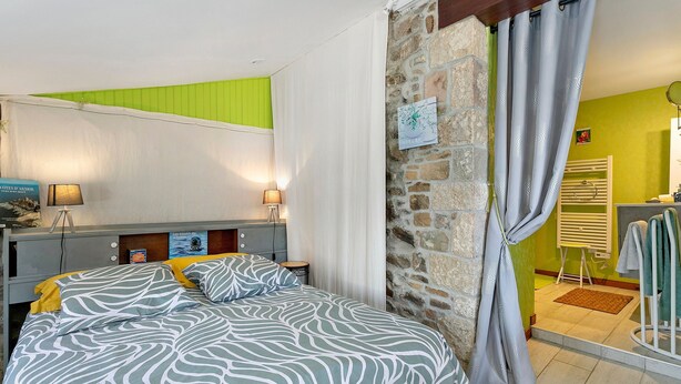 A bedroom with a bed, bedside lamps, a green wall, a stone wall, and a bathroom with a towel rack. - View of Château de la Roche-Jagu, Nordic Bath Relaxation Area – Guest Room (Ploëzal)
