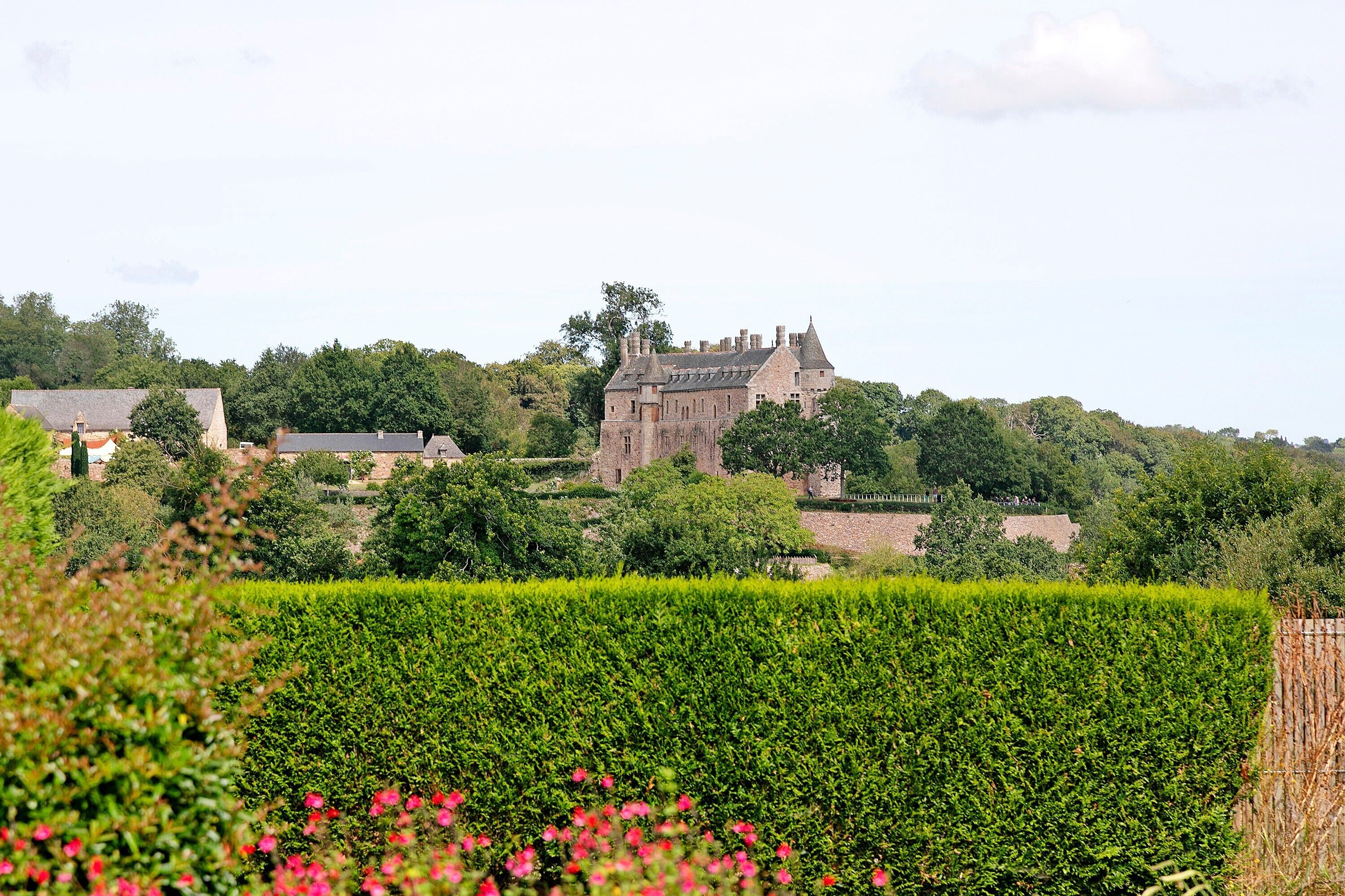 A historic stone building with a prominent tower, surrounded by lush greenery and a well-maintained hedge.