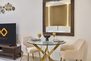 A modern dining area with a glass table, two chairs, and a television displaying a logo. - Stylish Studio with Dubai Canal & Skyline Views (Dubai)