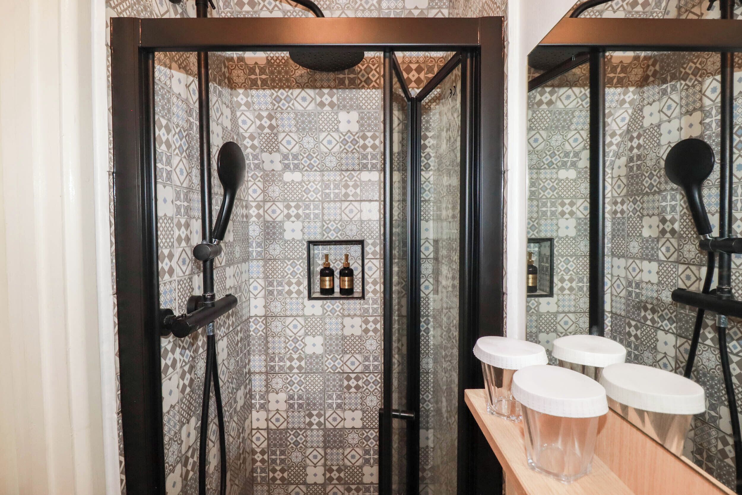 Zona de ducha con azulejos estampados, un espejo y una repisa con botellas y vasos.