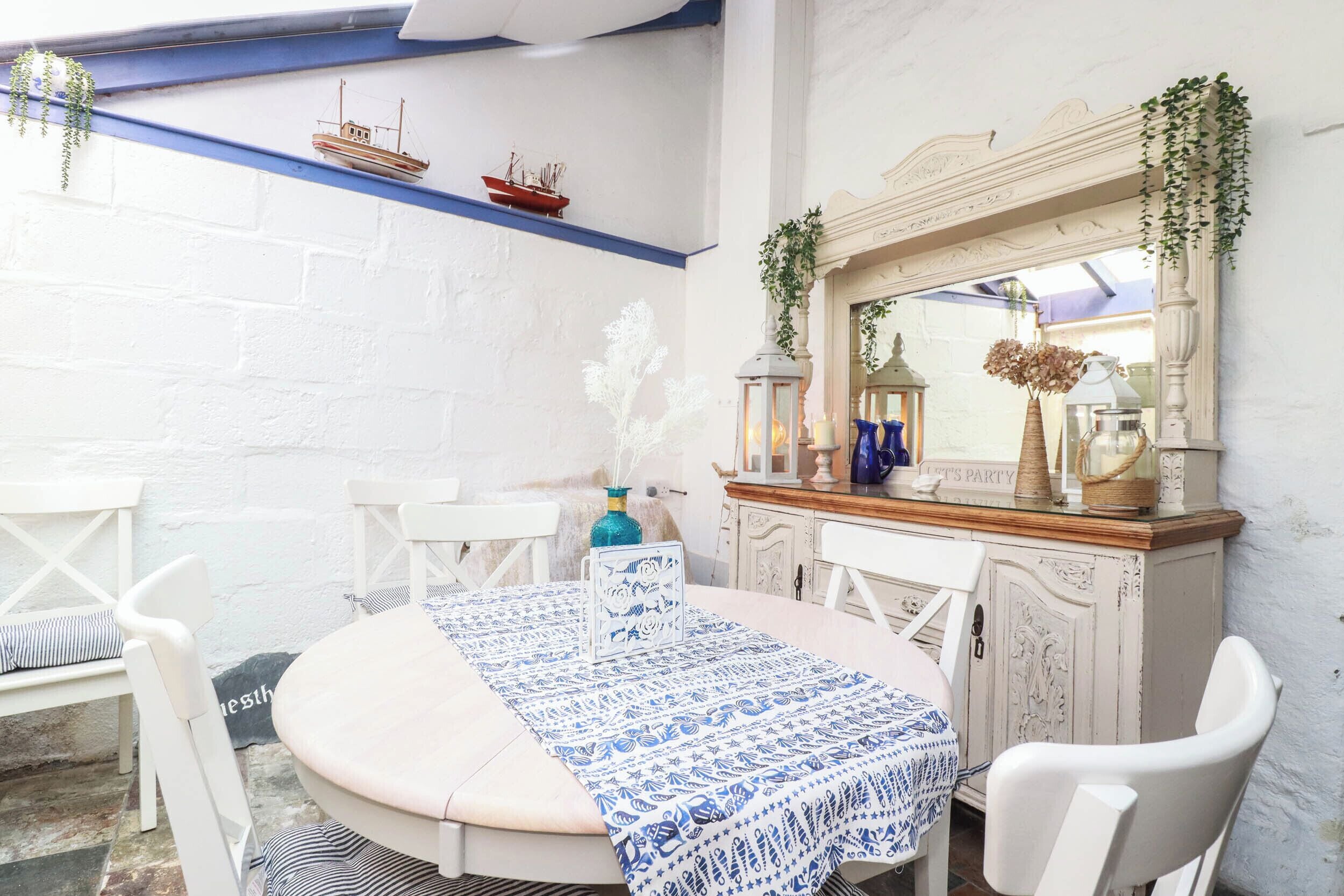 Un comedor con una mesa redonda, sillas blancas, mantel de tela azul estampada y un espejo decorativo sobre la mesa.